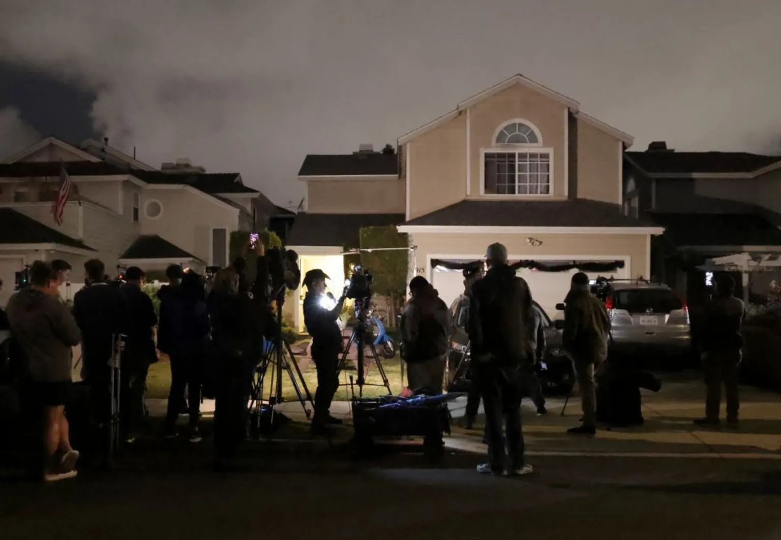 Media crews fill the street in front of suburban homes. It's dark and you can see the silhouettes of journalists and there are bright lights of TV crews illuminating their faces.