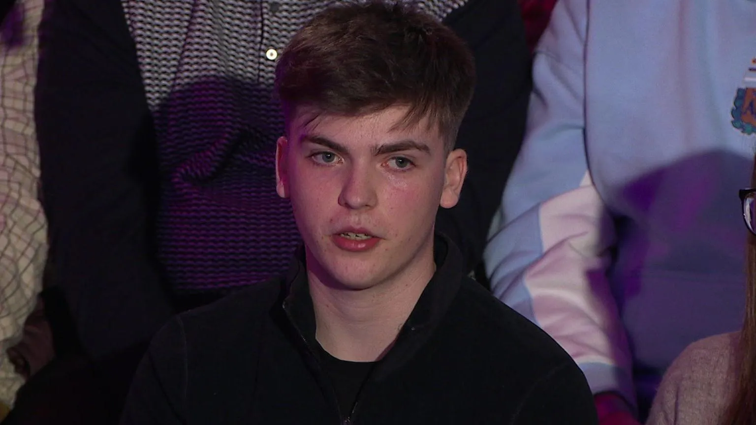 A man in a black top in an audience. He is the only one you can see.. but there are bodies of other men and women around him. It is a head and shoulders shot. He has dark brown, short hair.