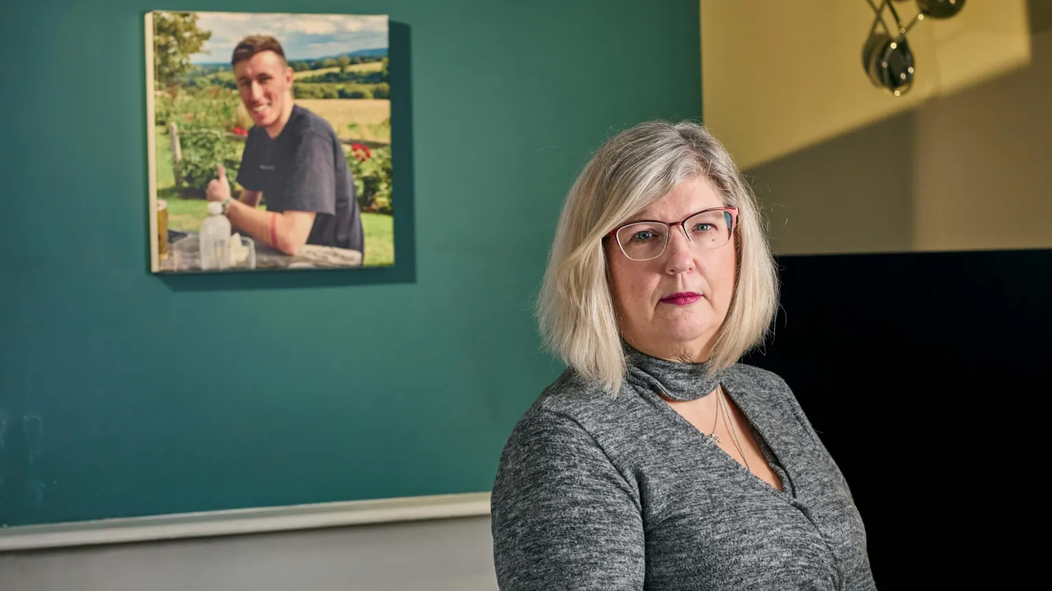 John Angerson A middle-age woman with blonde-greying hair stands to the right of the image. Behind her, hung on a green wall, is an canvas picture of his son, Bradley, who is smiling in front of a green fields