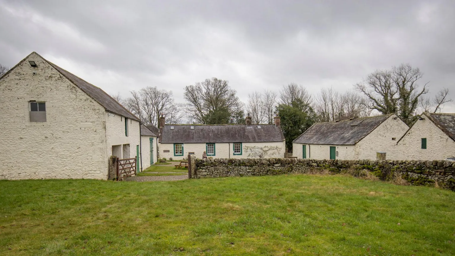 Mike Bolam A photograph of the farmstead. Tired-looking off-white buildings surround a courtyard. They are behind a dry stone wall and in the foreground is a large patch of grass. 