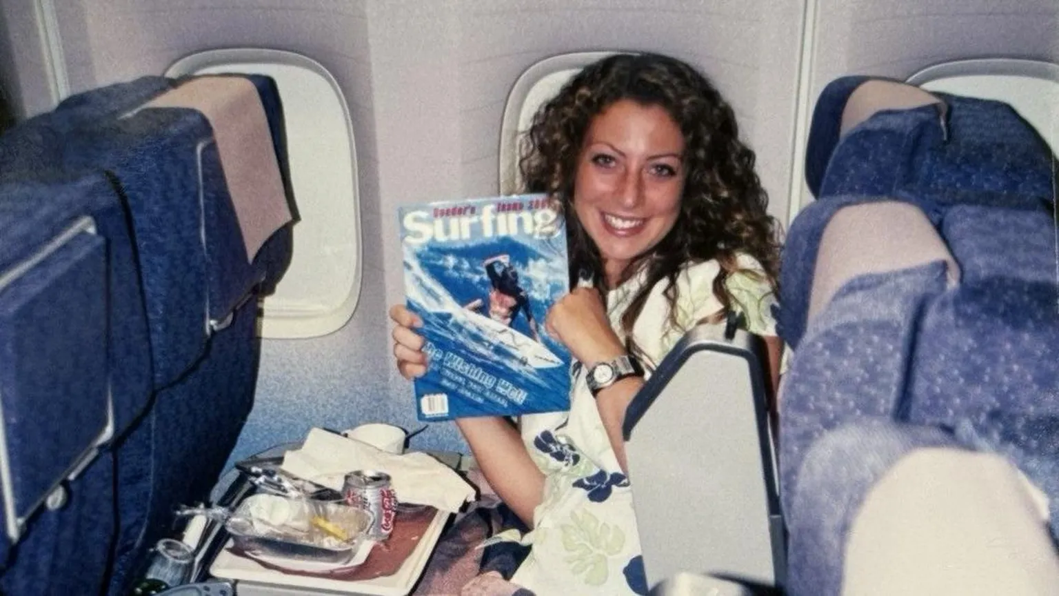 A young woman in a white T-shirt with long, curly auburn hair sits in a plane seat with a tray of plane food in front of her. She is smiling at the camera and holding up a surfing magazine. 