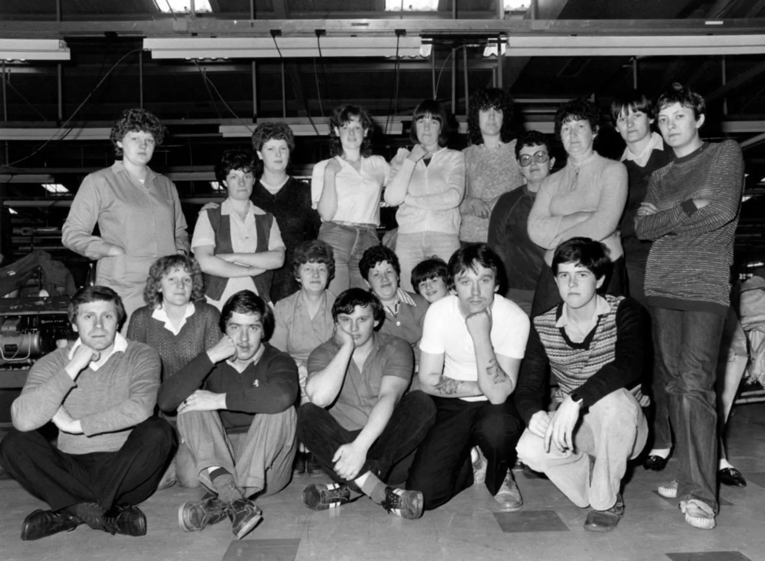 Mirrorpix via A black and white shot of factory workers in the 1980s. Men and women are pictured in three rows, those at the back are standing and those at the front are sitting.