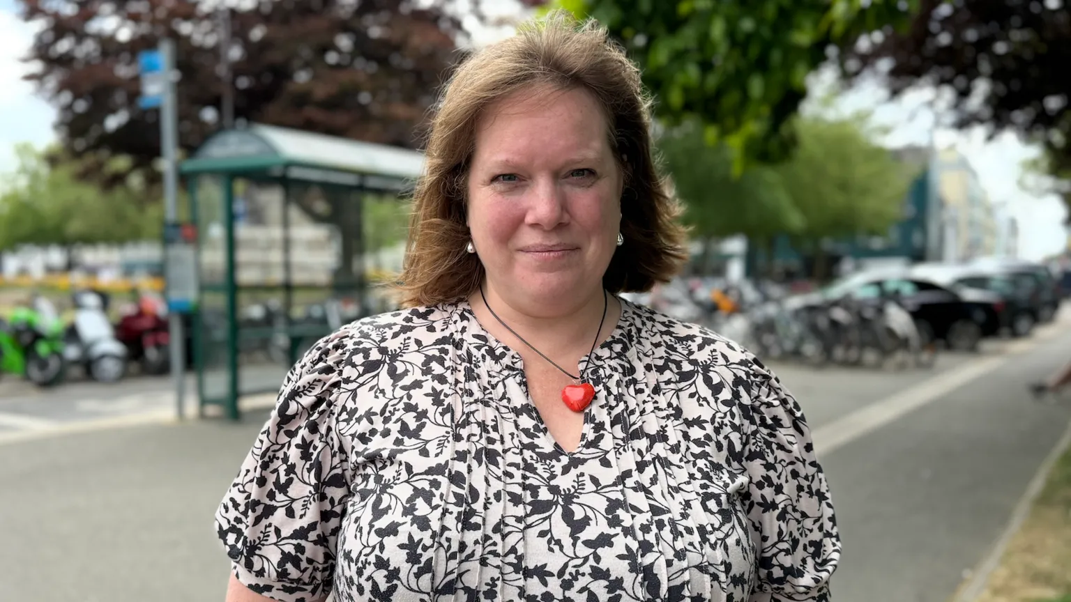 Kate Huntington is wearing a black-and-white patterned top and a red heart-shaped necklace. She is standing outdoors by a roadside, with a bus shelter, parked bicycles and trees visible in the background.