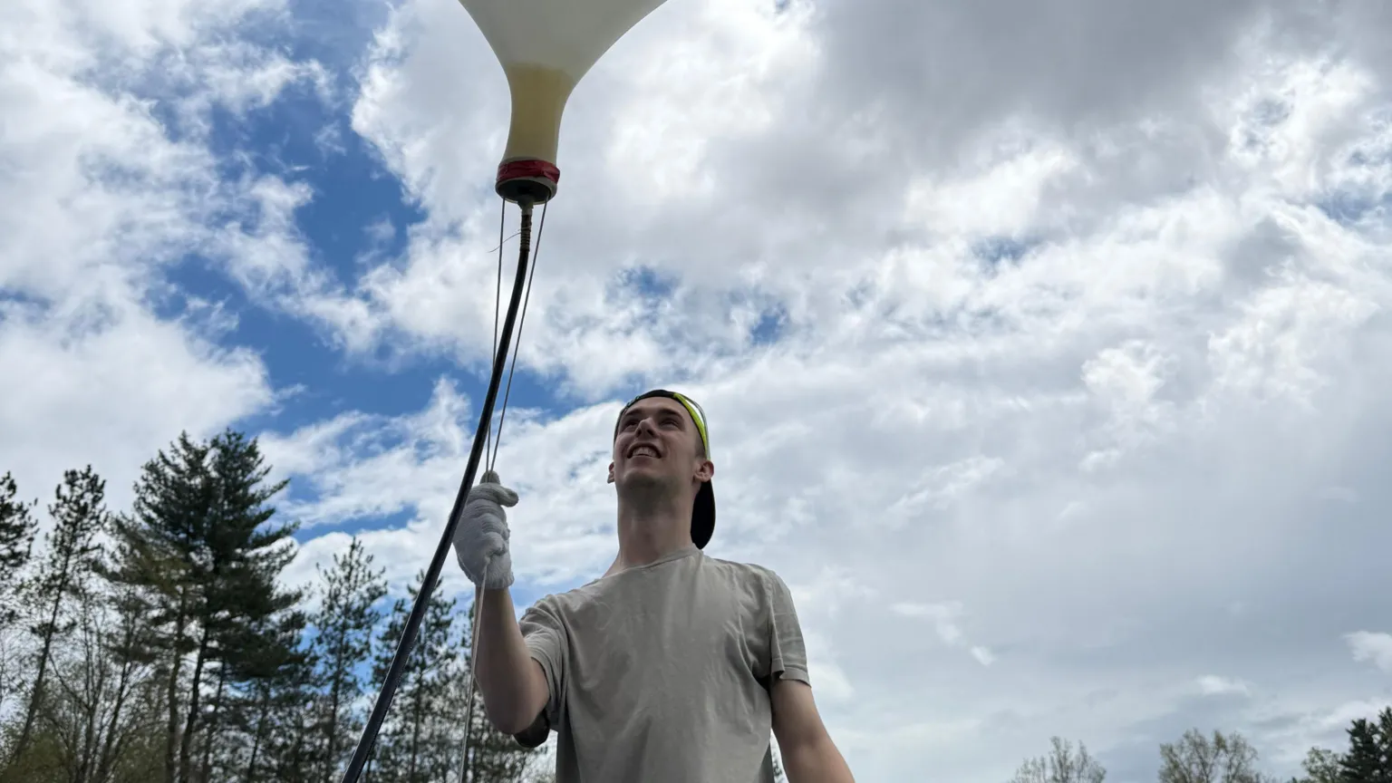Tom Liggett A white male in his twenties wearing a backwards baseball cap, t-shirt and white gloves, standing in a tree-lined field, holding a chord attached to a large inflated helium balloon, which is tethered to the ground before being released