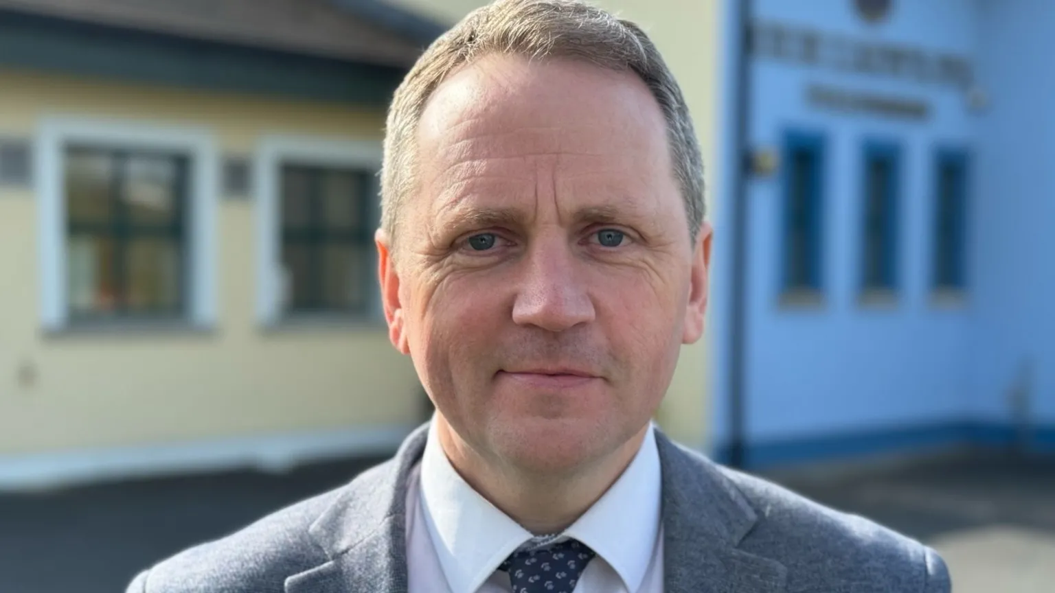 A man, with short brown hair, wearing a white shirt, navy tie and grey suit. There is a blue and yellow building behind him. 