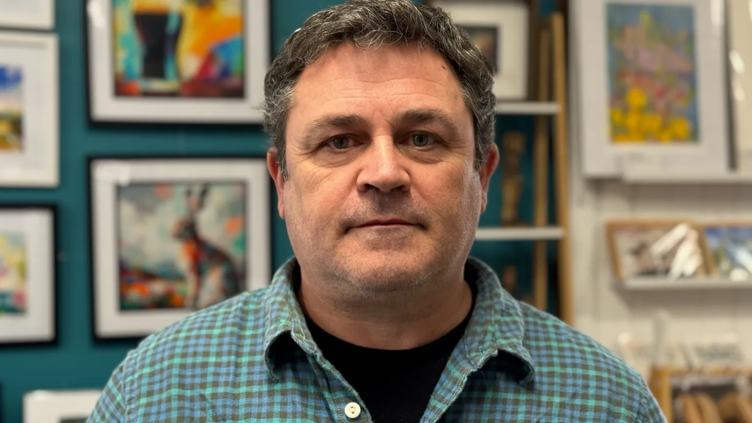 A man wearing a green, blue and navy check shirt and a black tshirt. He has short black and grey hair. There is a wall full of framed images behind him. 