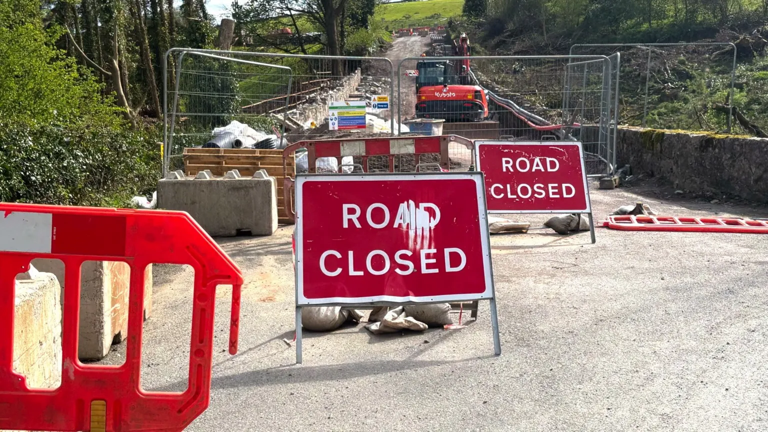 A road, closed off for construction. There are two red road closed signs and a digger behind the signs on the road. 