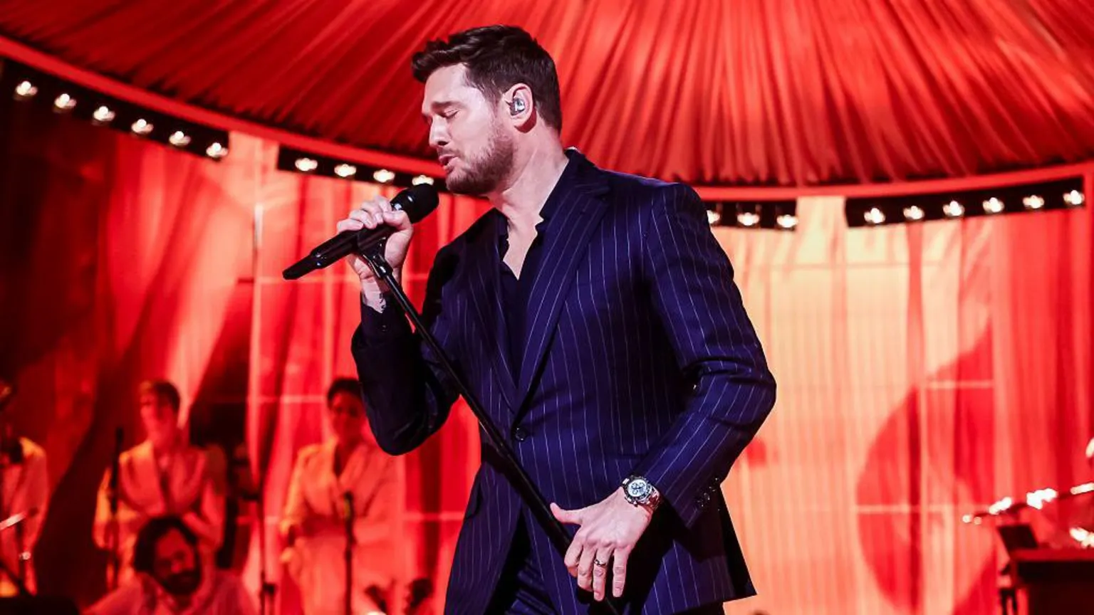  Michael Bublé singing into a microphone with red lighting. He is wearing a dark blue pin-stripe suit and watch.