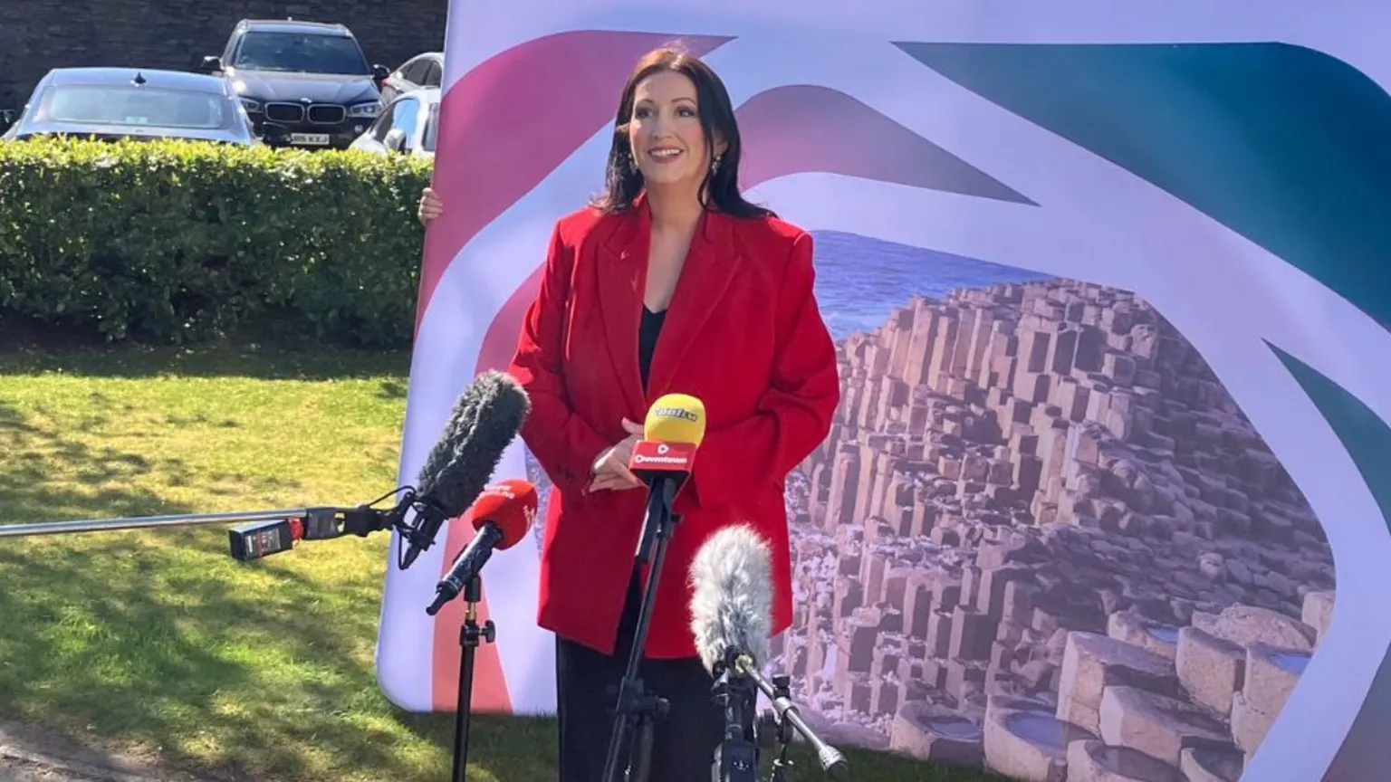 Shows a woman with long dark hair in red jacket and black dress standing in front of mics
