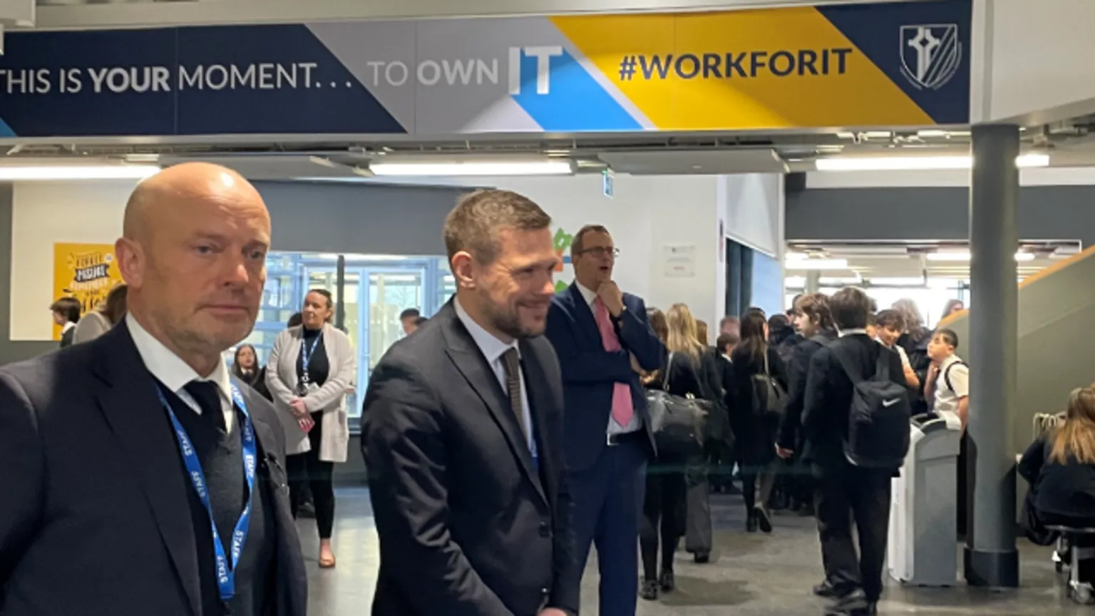 Three men in suits and ties standing in a busy area in a school with lots of school passing pupils. 
