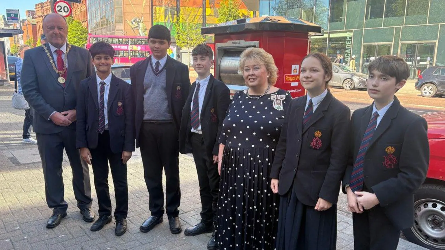 Four boys and a girl from Belfast Royal Academy, in dark school uniform, in front of the new red postbox featuring the yellow cypher of King Charles III. To their left is a tall man in a grey suit with a red tie and a gold chain of office around his neck. Also in the picture, third from the right, is a blonde-haired woman in a navy and white polka dot dress.