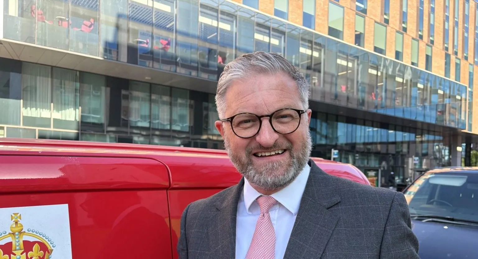 A man with, grey hair and dark glasses and a grey beard, wearing grey suit, white shirt and pink tie, beside a red Royal Mail van in Belfast's York Street