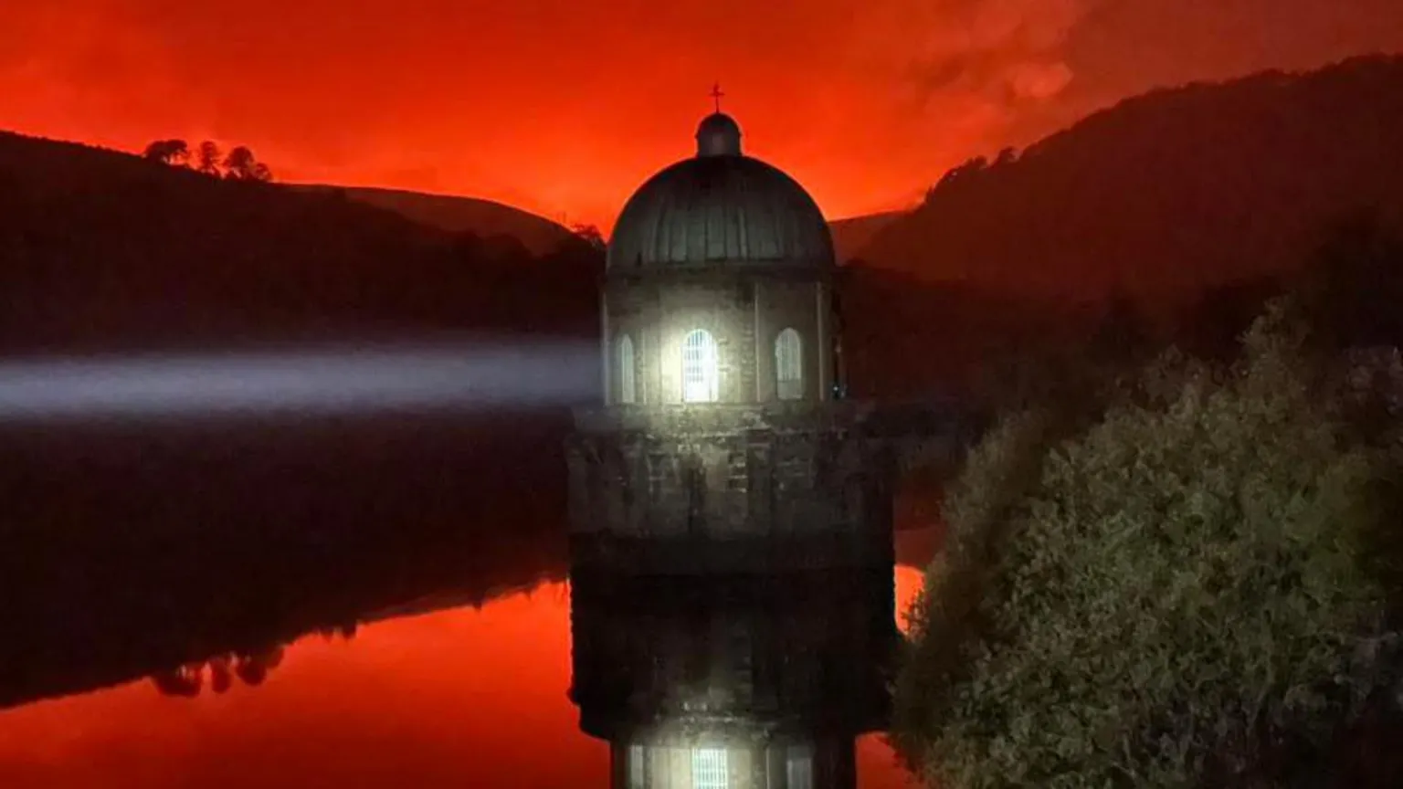 MWWFRS Rhayader A lighthouse is pictured in the water, shining a light. Above it is a red sky, lit up by wildfires in the distance. The red colour is also reflected in the water.