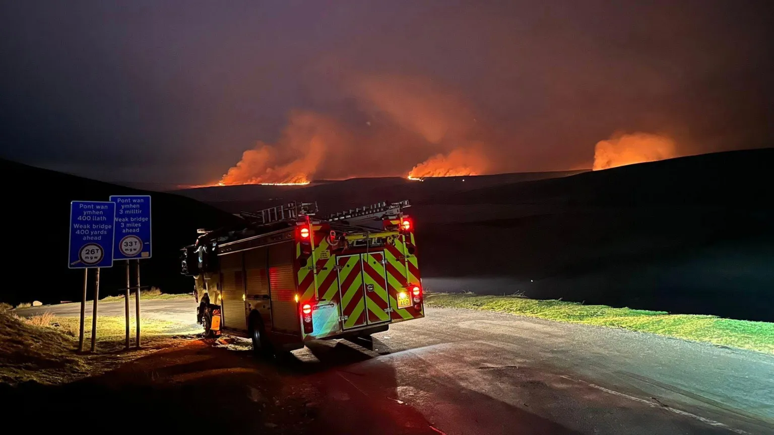 MWWFRS Rhayader A fire engine is pictured on a road under the night sky. Wildfires have lit up the dark with several spots pictured wit large amounts of smoke.