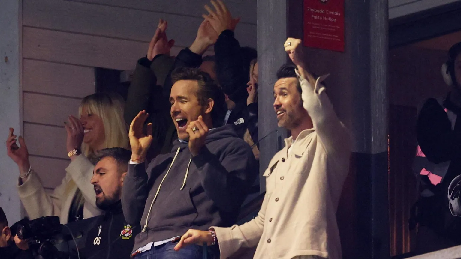 Action Images/ Rob Mac and Ryan Reynolds celebrating a goal at a Wrexham football match. Rob is holding his fist in the air and Ryan is smiling with his mouth open, mid applause.