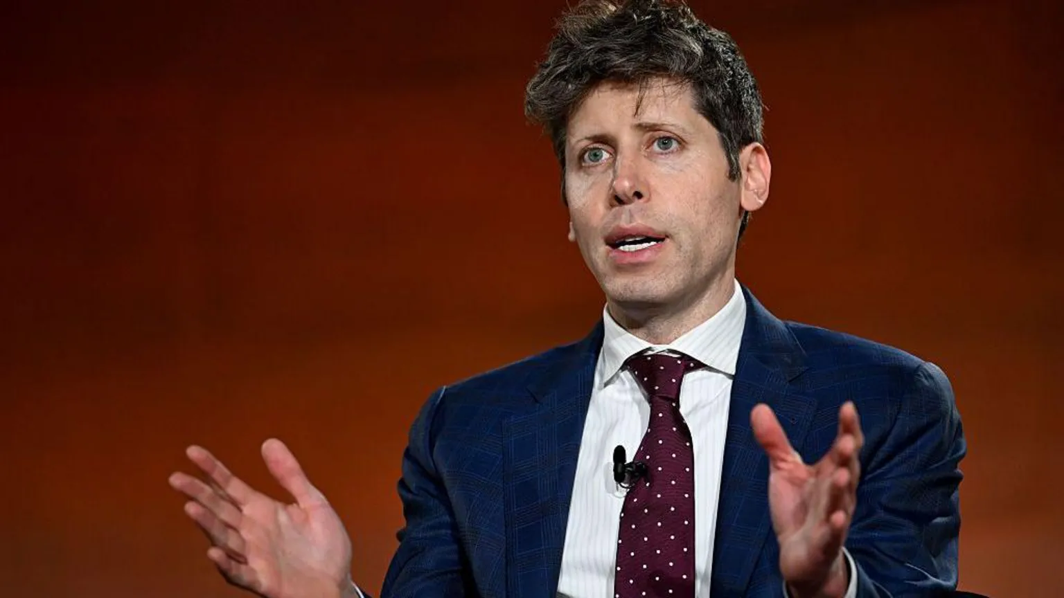 Bloomberg via An image of Sam Altman speaking. He has blue eyes and short dark brown hair, and is wearing a navy blue blazer with a white shirt underneath and a burgundy tie with small white polka dots. He is gesturing with both his hands addressing a crowd off camera, behind him is a blurred copper background 