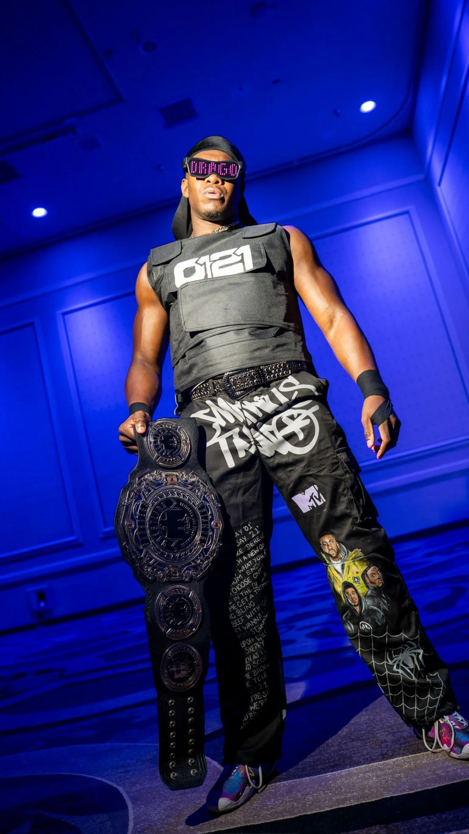 Jordan Lees Pro wrestler DeReiss Gordon, a muscular man poses confidently in a room bathed in blue lighting. He wears sunglasses displaying 