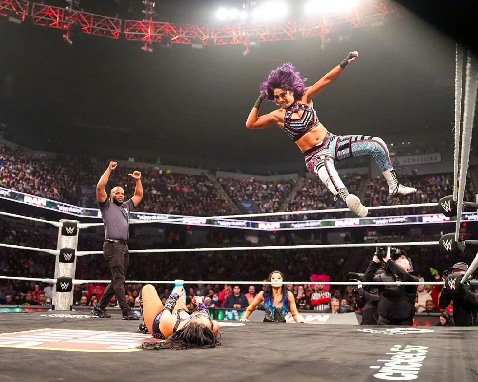  Bayley leaps from the top turnbuckle in a wrestling ring, delivering an elbow drop to a prone opponent.