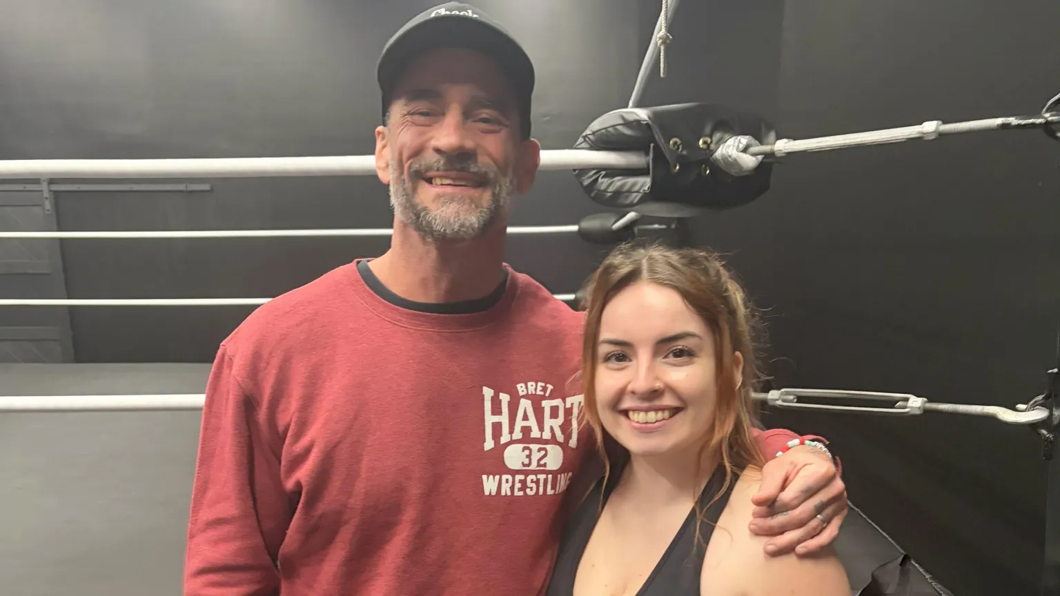 Natasha Swann CM Punk and Natasha Swann standing in a gym, just outside a wrestling ring. He is wearing a light pink top. Natasha is wearing black work out gear.
