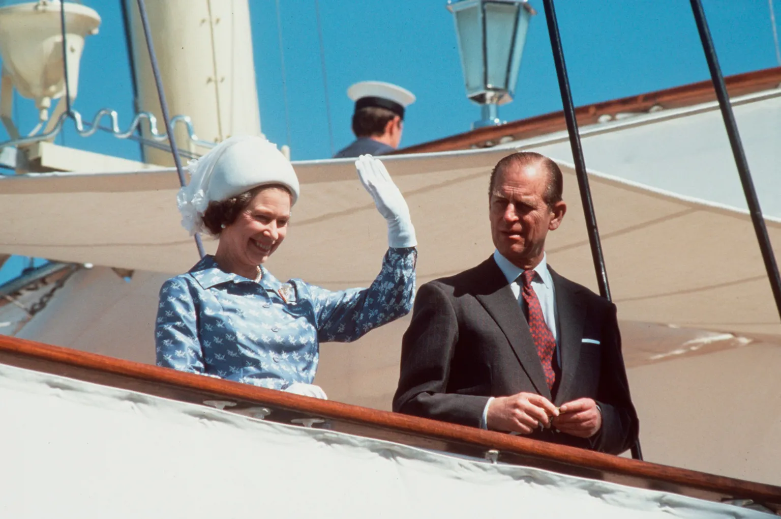  A man and a woman stand together on a boat. The women is wearing a blue dress, a white hat and white gloves. She is waving her hand. The man is in a black suit with a red tie.
