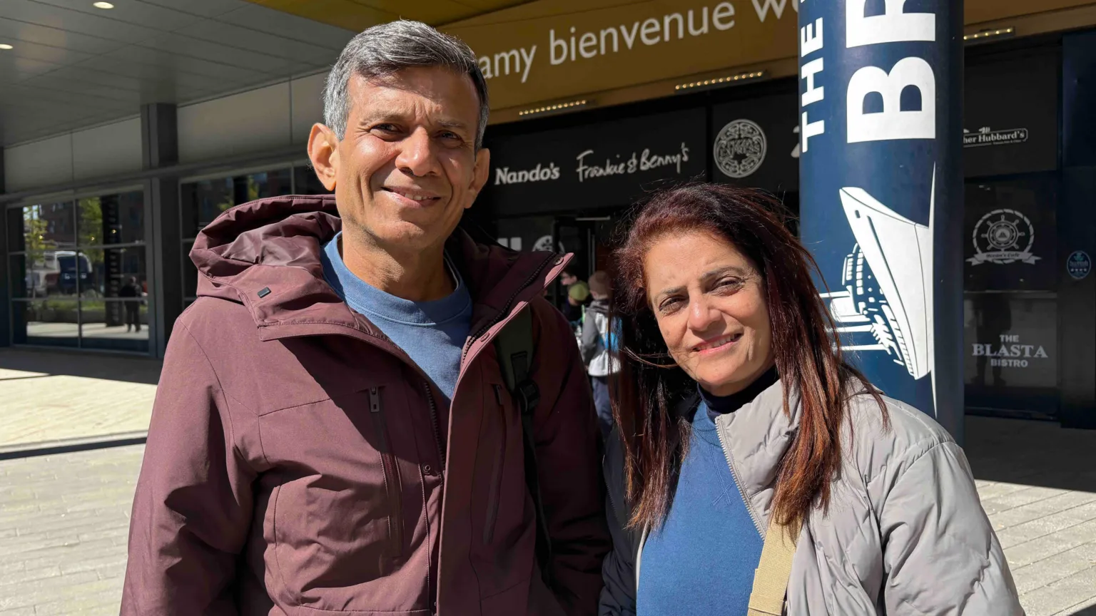 A man and a woman are stood smiling at the camera. The man has grey hair and is in a red jacket and has a blue shirt on underneath. The woman has red hair and is wearing a grey jacket with a blue jumper underneath.