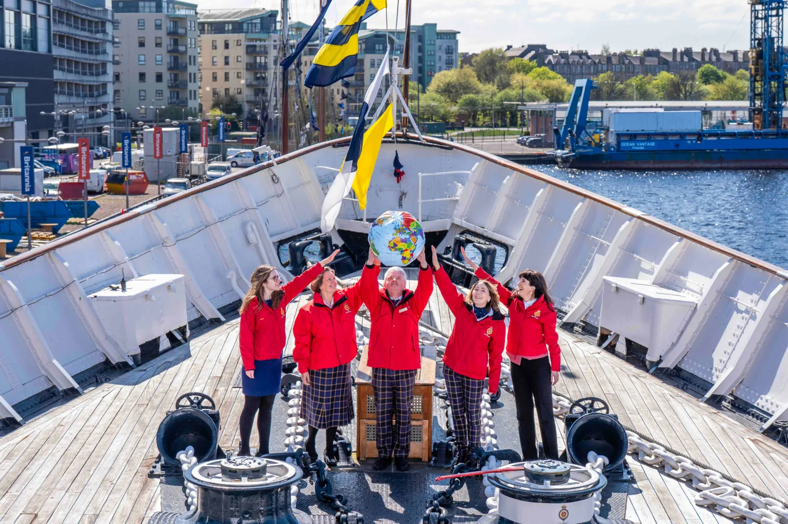  A group of four people hold a blow-up globe. They are stood on a boat and are all in red jackets and tartan bottoms.