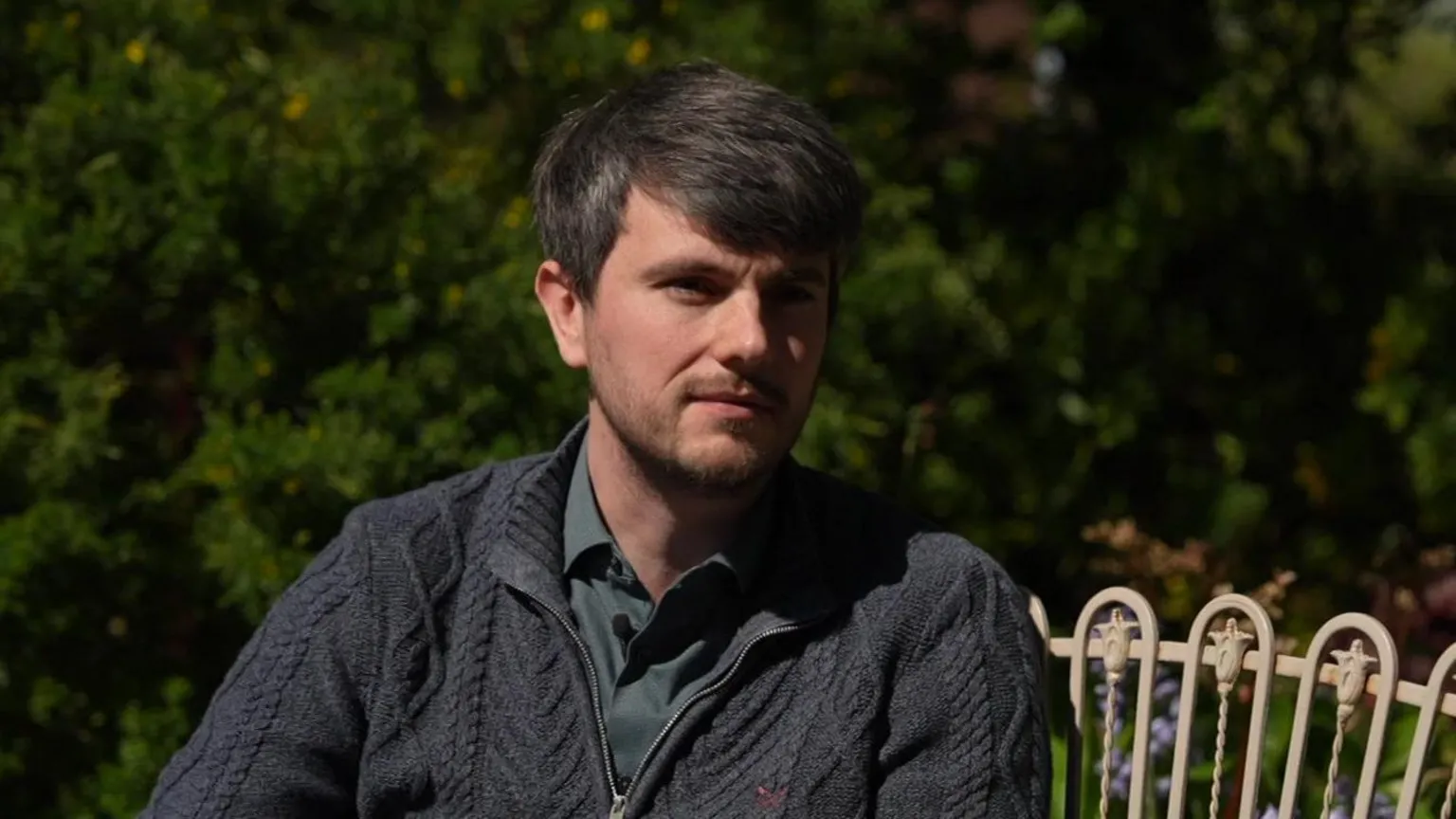 A man in a cable-knit jumper and shirt, sitting on a bench. There are trees and shrubbery behind him.