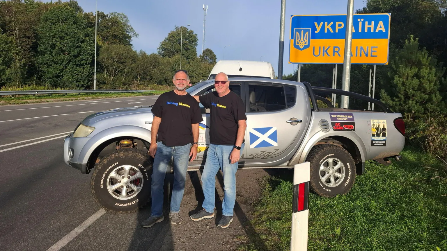 Steve Eccleshall Two men stood in front of a pick-up truck next to a sign marking the Ukrainian border