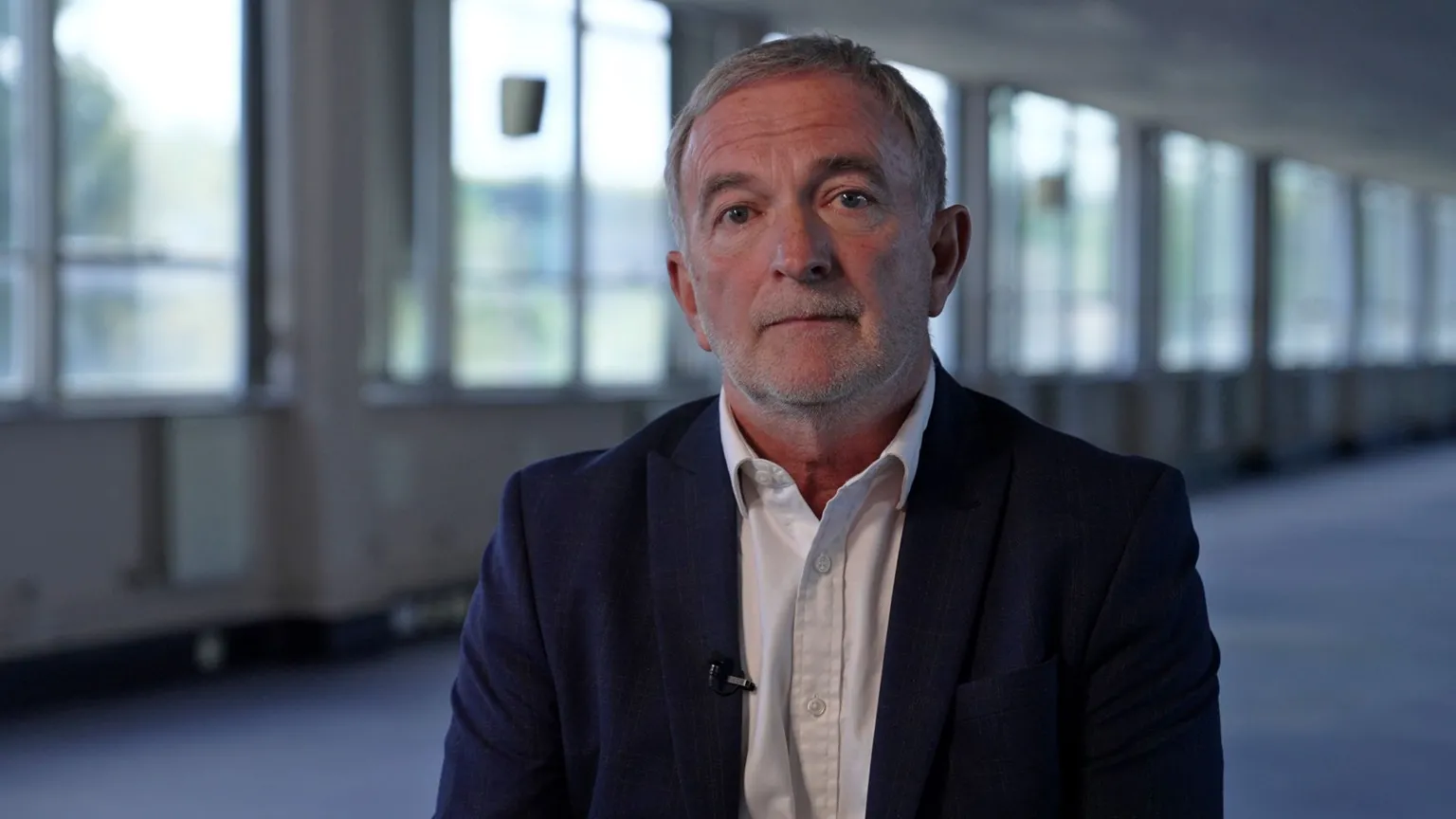 Andrew wearing a dark blazer and light shirt, seated indoors in a modern office corridor with large windows and soft natural light in the background.