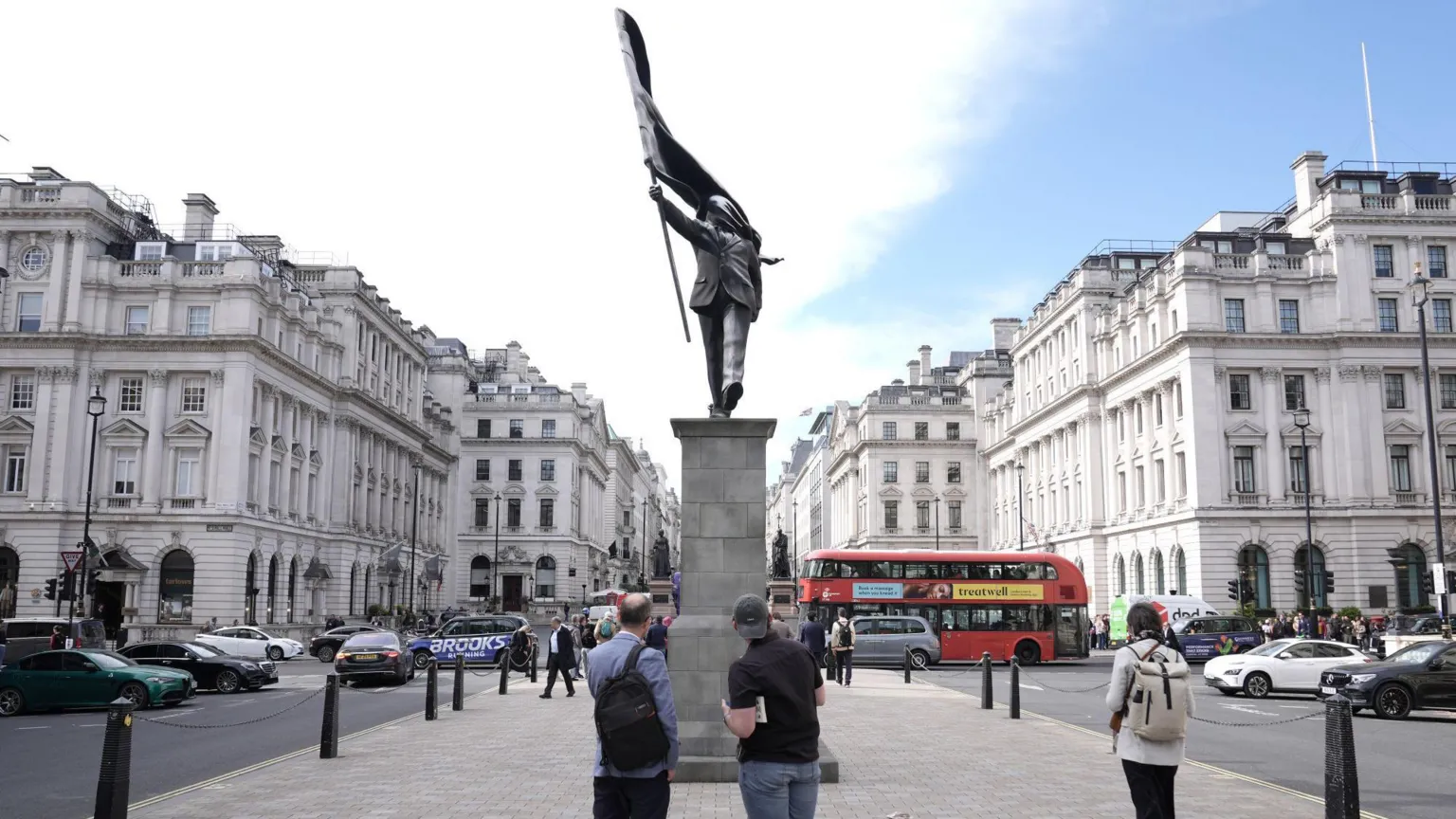  The statue of a man marching while holding a flag seen on a plinth front view, with three people stopping to look
