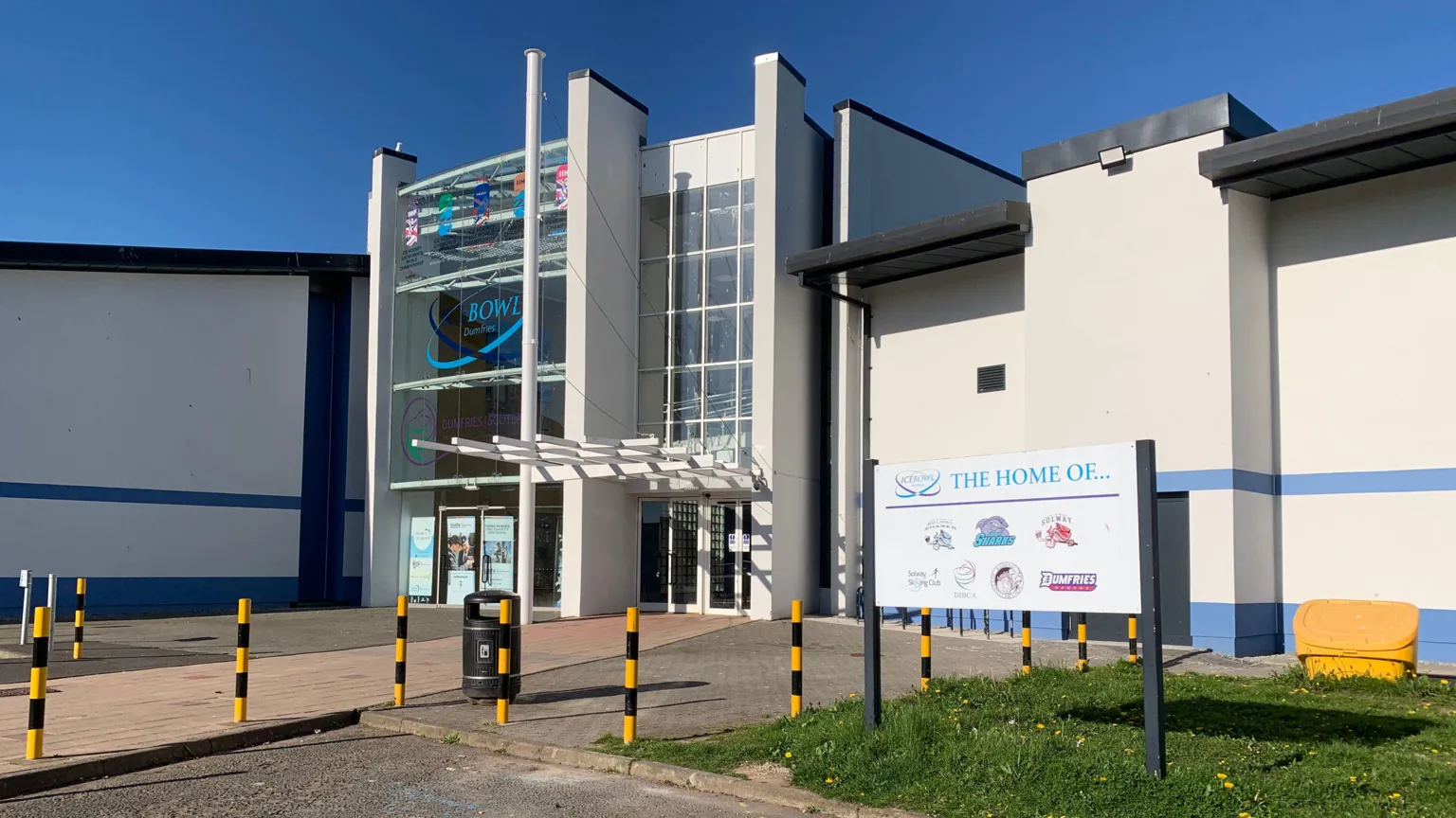 The Dumfries Ice Bowl with a white and blue facade and glass entrance way. There are black and yellow bollards outside, a sign saying what teams play there and a black bin.