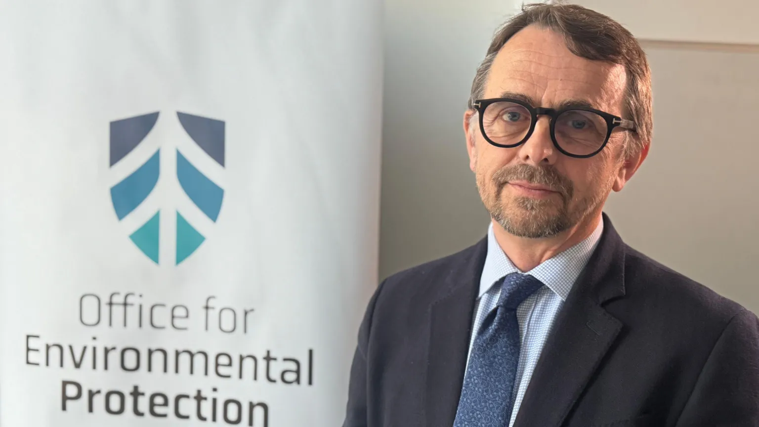Robbie McDonald is wearing a navy blazer with blue shirt and tie. He has black glasses on. The background is a white poster for the Office For Environmental Protection. 