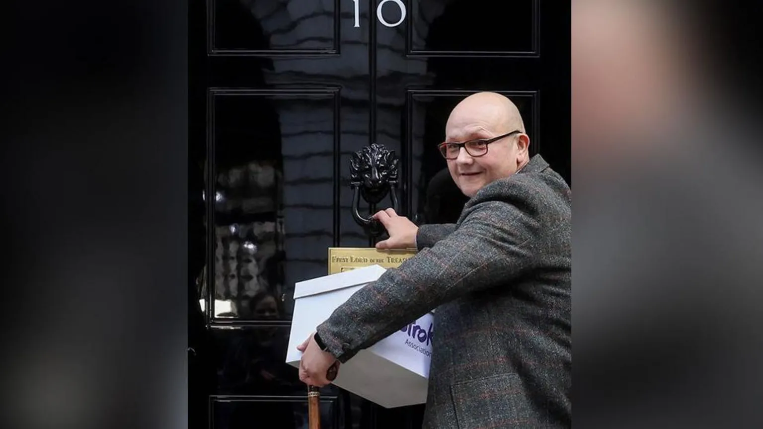 Stroke Association/PA Wire Campaigner Phil Woodford who is bald and wearing glasses and a tweed blazer knocks on the door of No 10 Downing Street with a box of signatures. He is smiling.