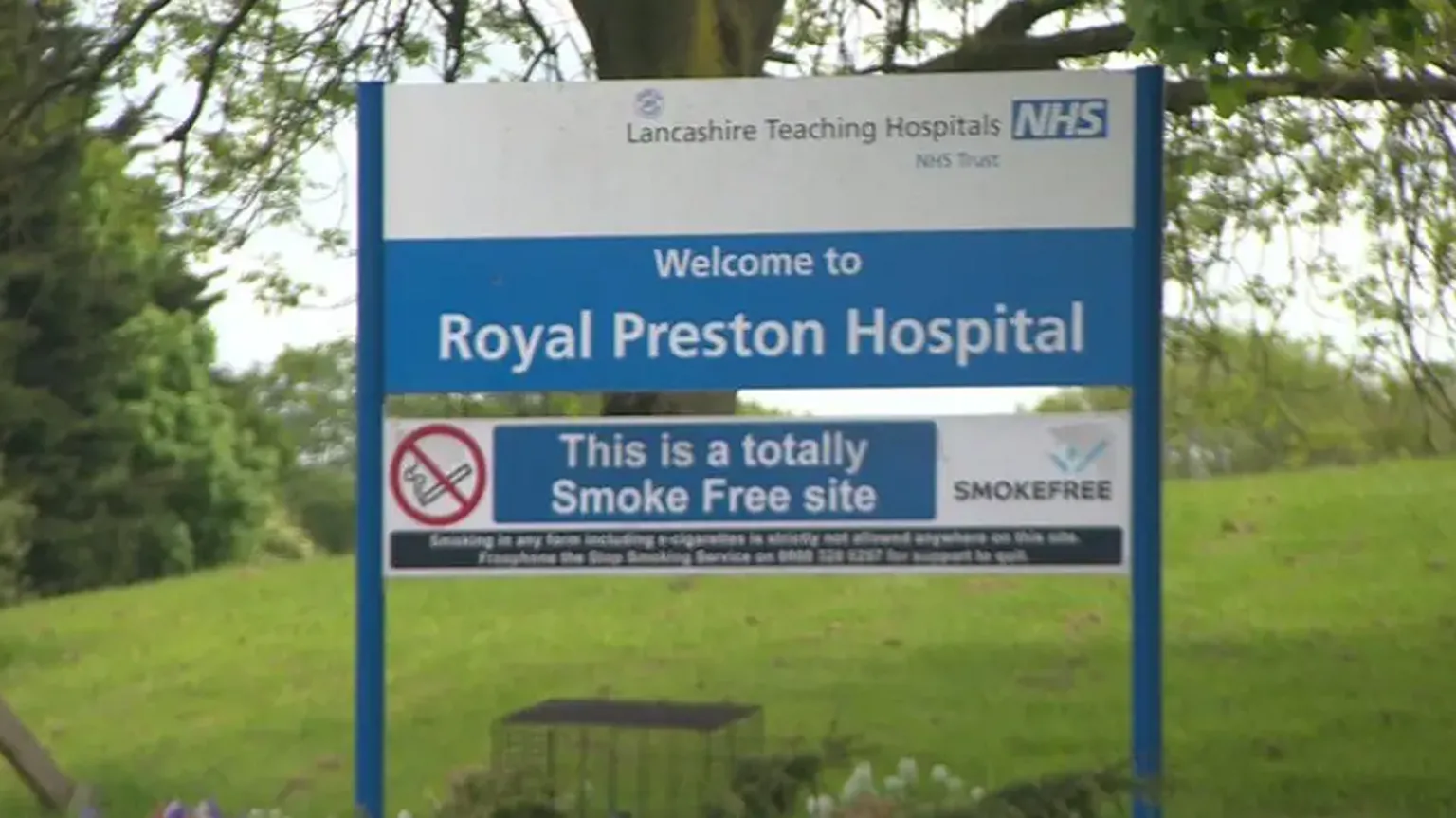 A sign saying 'Welcome to the Royal Preston Hospital' and 'Lancashire Teaching Hospitals NHS Trust' is on a steep bank of grass, with a plant beneath it and trees nearby.