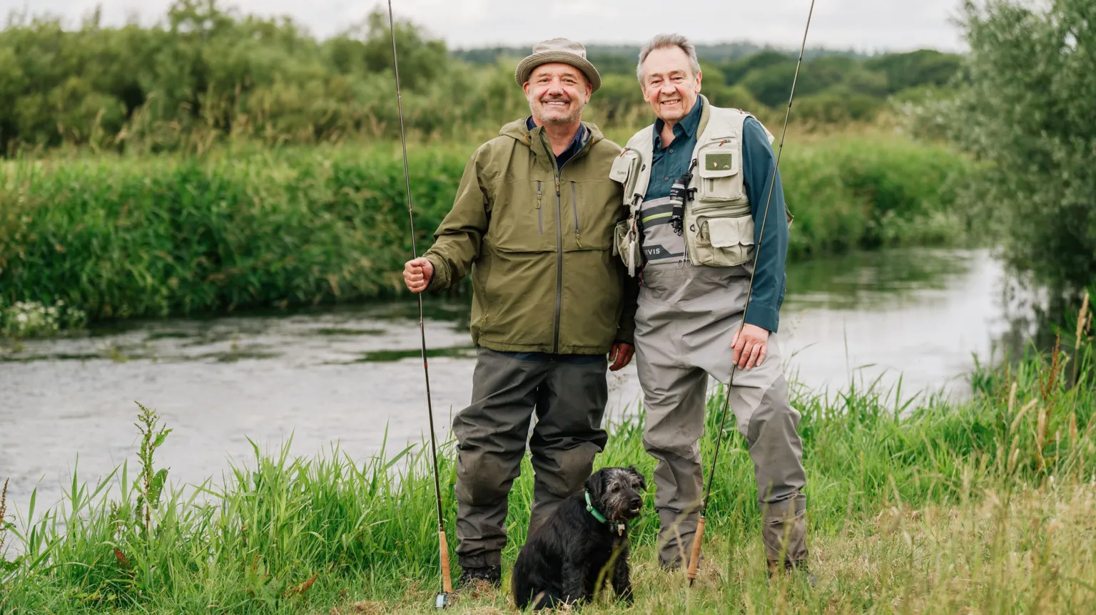 BBC/Owl Power/Sam Gibson Bob Mortimer and Paul Whitehouse pose for a photo, holding fishing lines with standing by a river, with small black dog Ted in front of them.