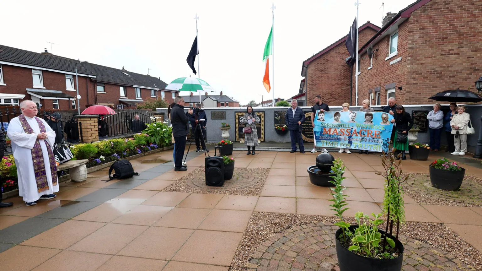 Pacemaker People gathered in a paved courtyard. A priest in on the left and people in the right corner are holding up a banner reading: 
