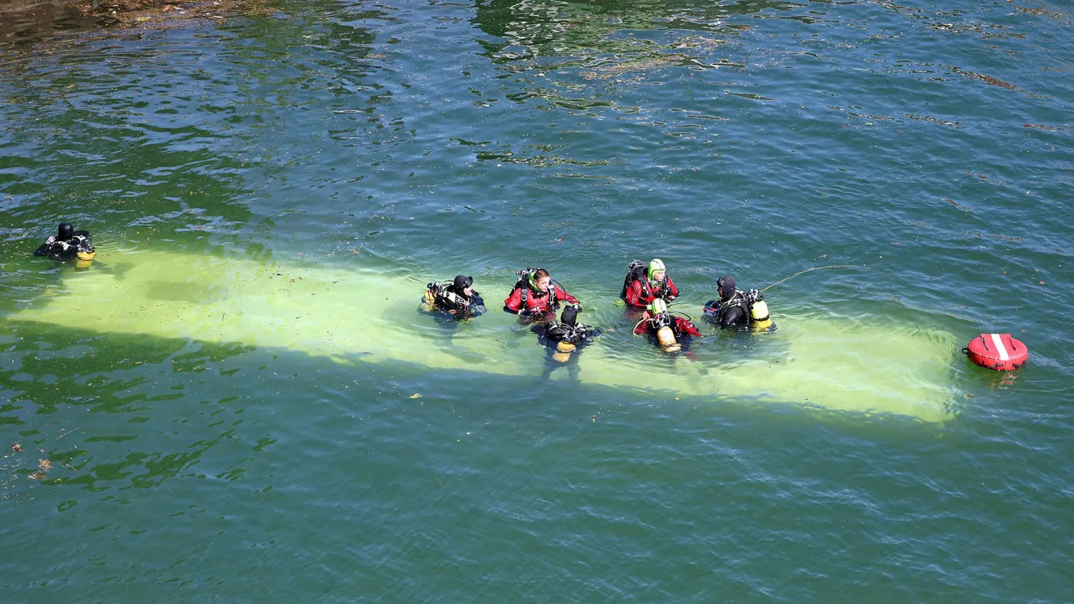 The roof of the bus is visible under the river. Six people in wetsuits are sat atop it, and one more person is in the water on the left of the frame.