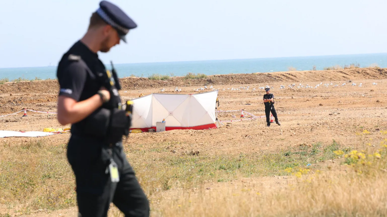 UKNIP A police officer stands in the foreground. In the background is a white police tent just in front of the beach. A police officer stands next to it.