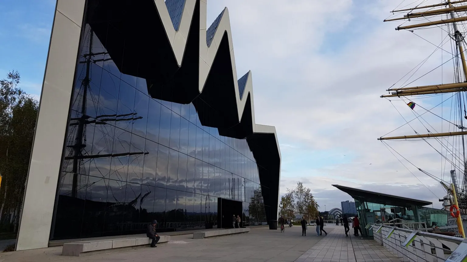  A large museum building, by the riverside. Most of the building's wall is glass, with a jagged concrete pattern at the top