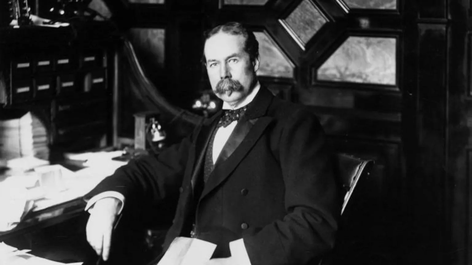  An old black and white photograph, showing Thomas Lipton - a man in a suit, with bow tie and with a bushy moustache, sitting at a desk