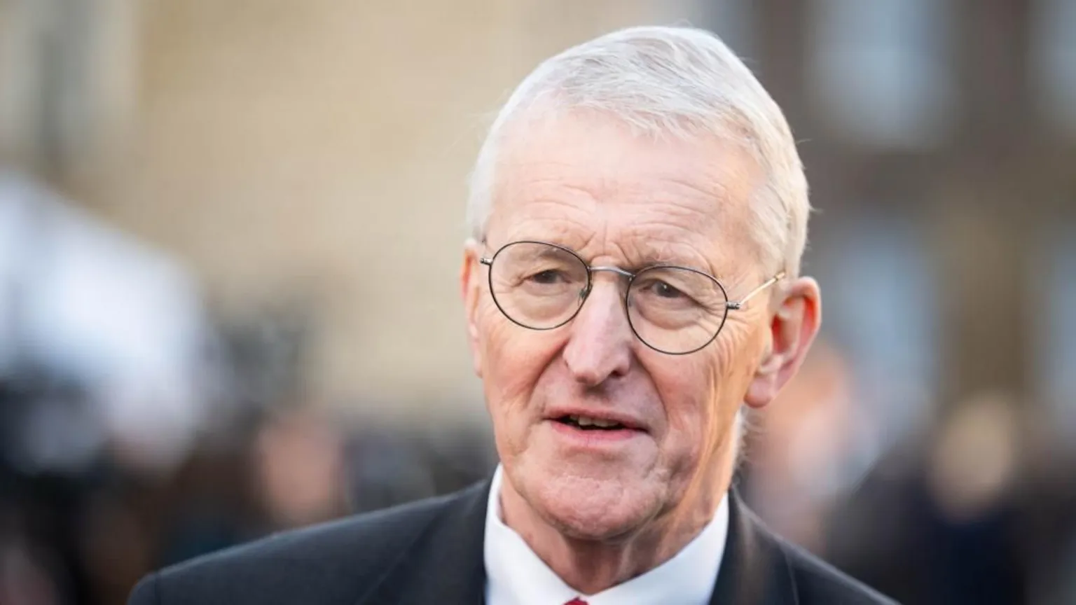  Hilary Benn is an older man with short white hair. He is wearing a dark suit, white shirt and round metal framed glasses.