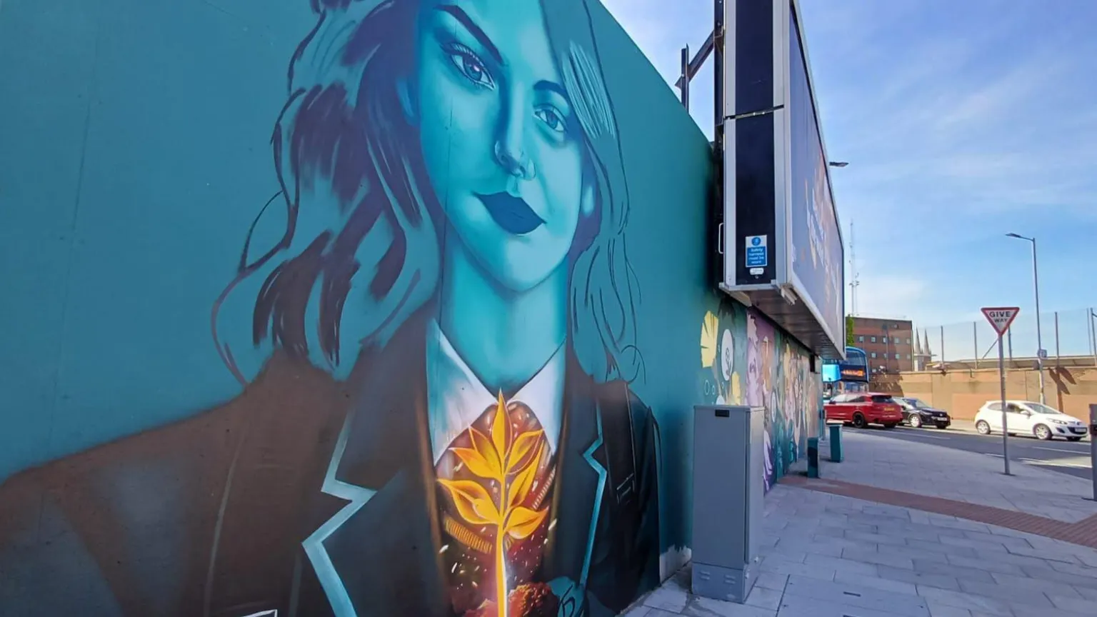 A mural of a teenage girl wearing her school blazer. The mural is mostly green and she's holding a gold leaf. Traffic is in the background. 