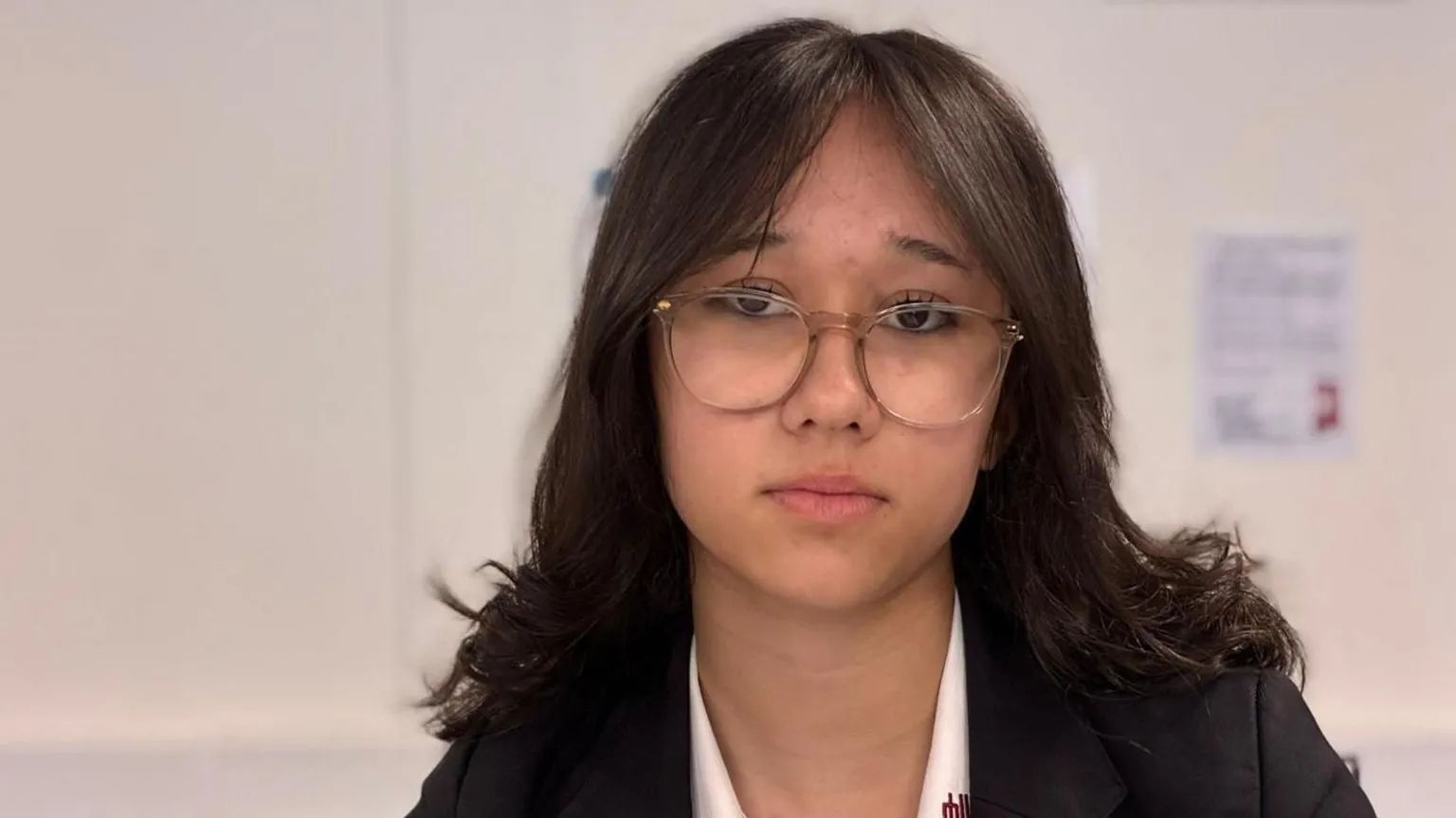 A teenage girl with shoulder-length dark hair and glasses. She is wearing a white shirt and black blazer and looking at the camera with a serious expression.