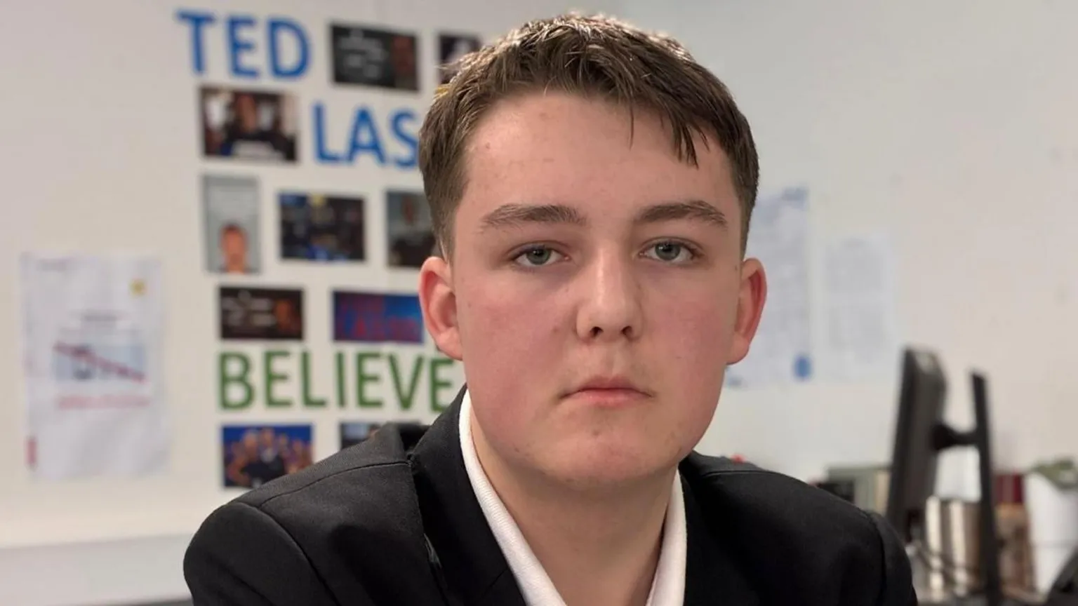 A boy with short brown hair. He is wearing a white shirt and black blazer and is looking at the camera with a serious expression.