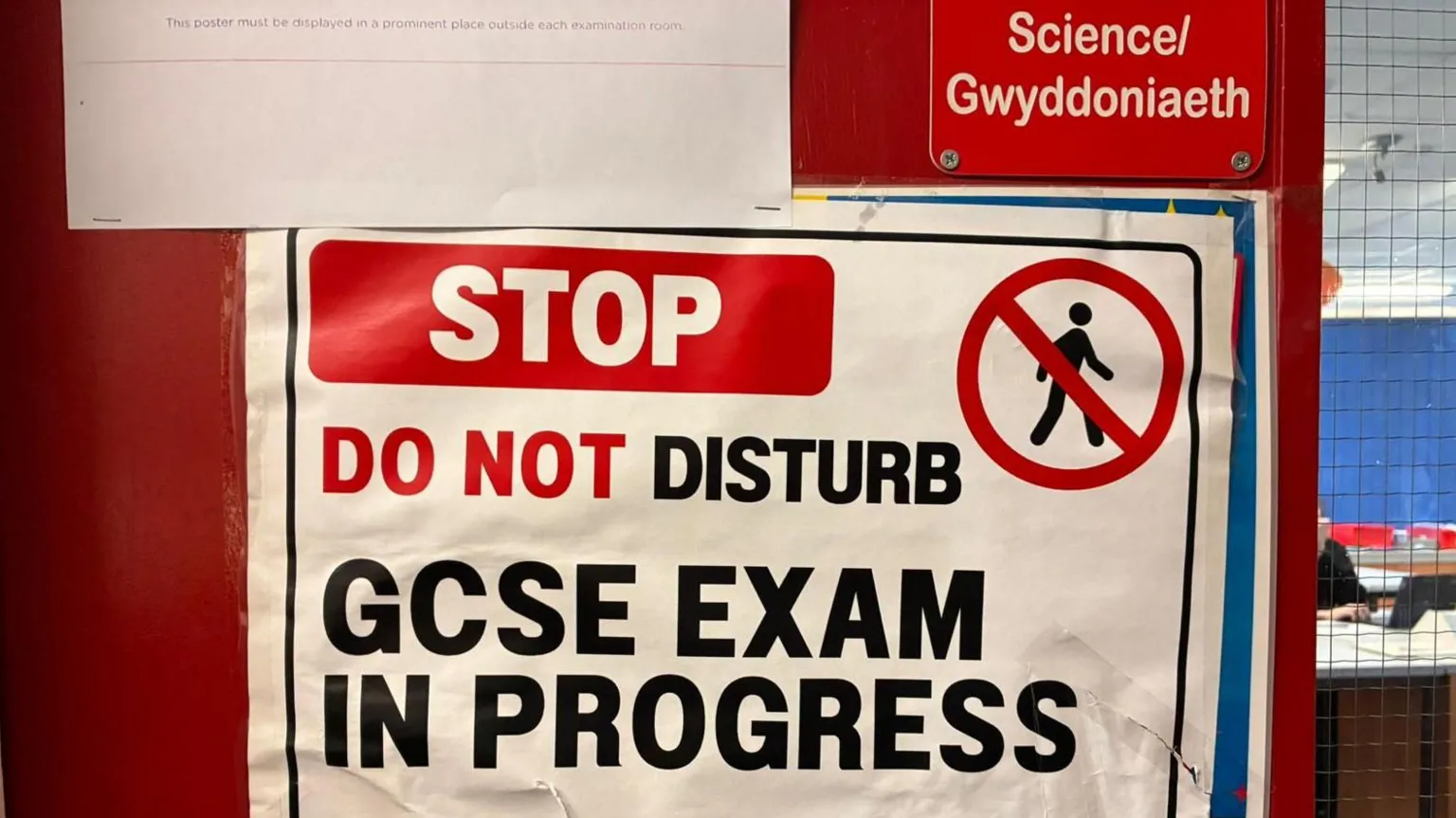 A paper sign pinned to a red door. The door has a sign saying Science and a small window which shows pupils at desks inside the room. The sign reads: 