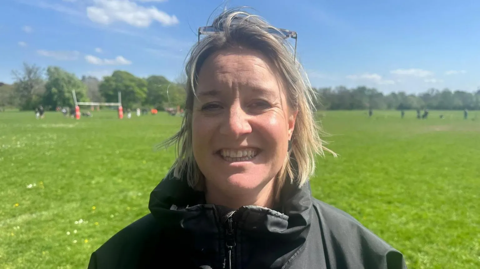A woman with short blonde haair wearing a black jacket and sunglasses on top of her head. She is stood in a rugby field with a match going on on the green grass behind her. It is a sunny day with blue sky. She is smiling and looking at the camera.