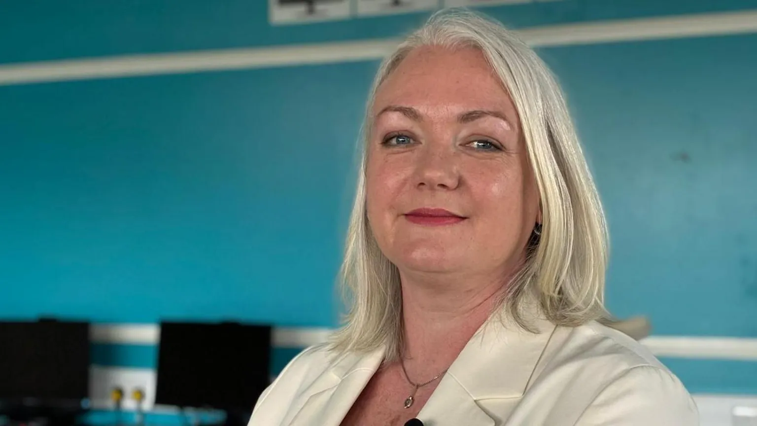 A woman with white grey shoulder-length hair. She is wearing a white blazer. She is stood side-on and looking at the camera with a small smile.