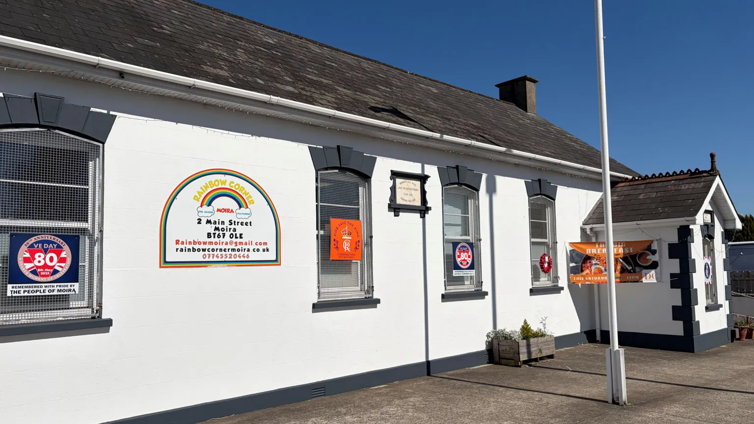 Deramore Orange Hall, a single-storey Victorian building painted white with a grey slate roof, to the right of the photo is a covered porch of the same construction and a flagpole stands in the centre of the image. Rainbow Corner's logo is on a sign in the centre of the wall.