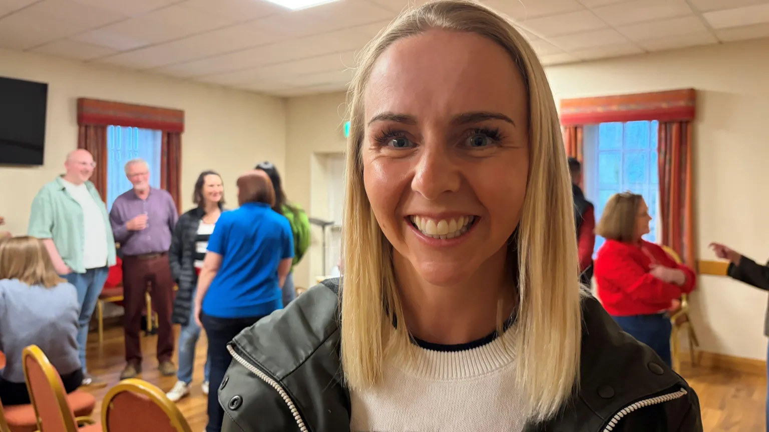 Laura Coney has shoulder length blonde hair and is wearing a white knitted sweater with a dark green raincoat over the top. She is standing in a hall, behind her various other people can be seen standing around speaking to each other.
