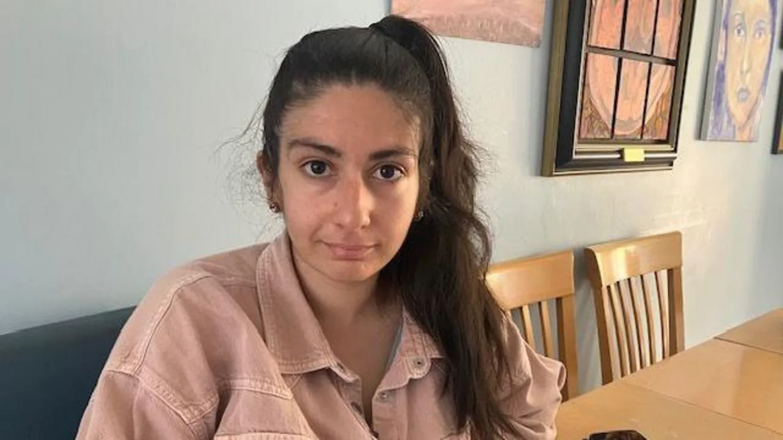 A young woman with long dark hair tied in a pony tail sits at a dining table with artwork on the wall next to her. 